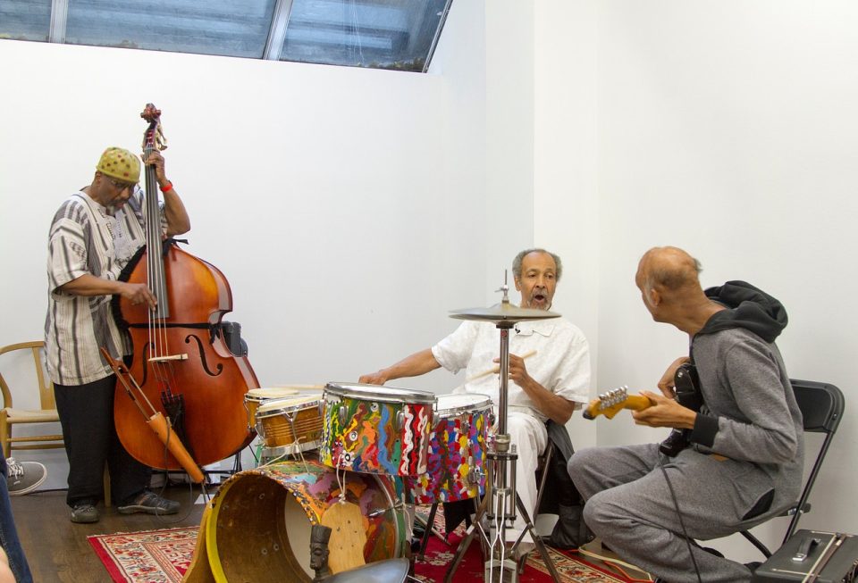 Three musicians in a room with a red carpet. The figure on the left is playing an upright bass, the center figure is seated behind a drum set, and the figure on the left holds a guitar with their back turned to the camera.