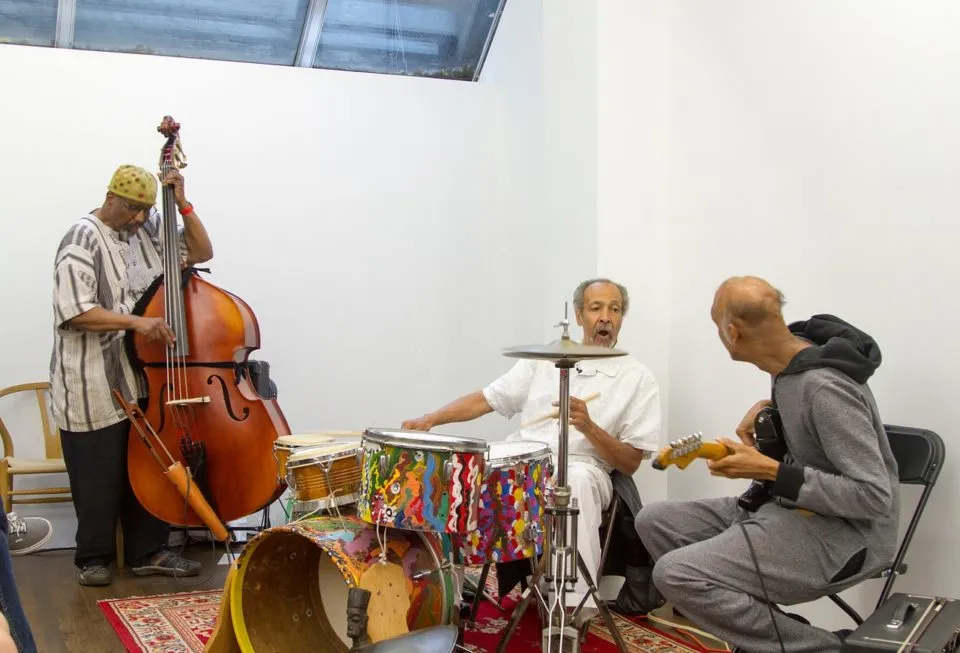 Three musicians in a room with a red carpet. The figure on the left is playing an upright bass, the center figure is seated behind a drum set, and the figure on the left holds a guitar with their back turned to the camera.